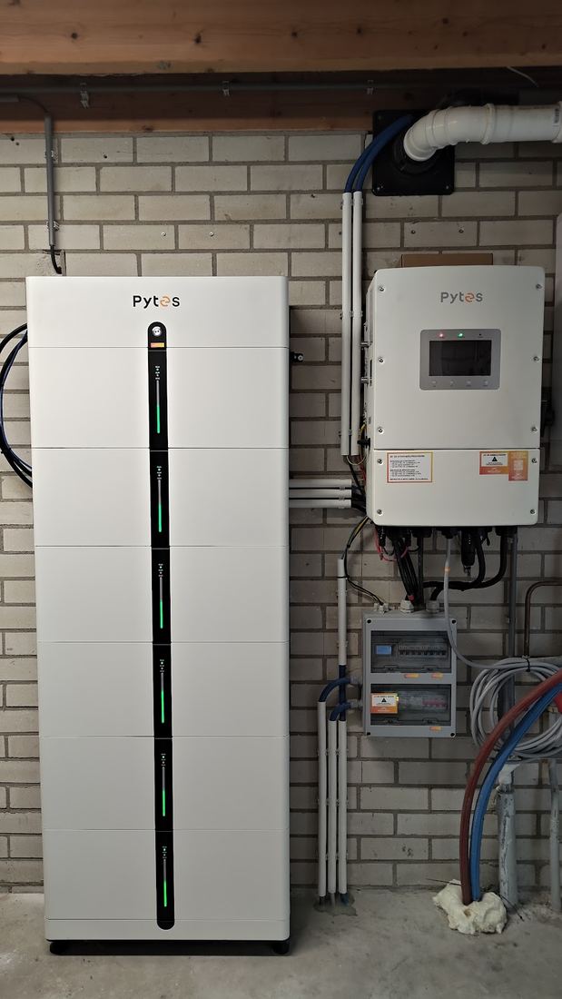 Een hoge witte accu met daarnaast de omvormer en een kleine groepenkast tegen een witte muur