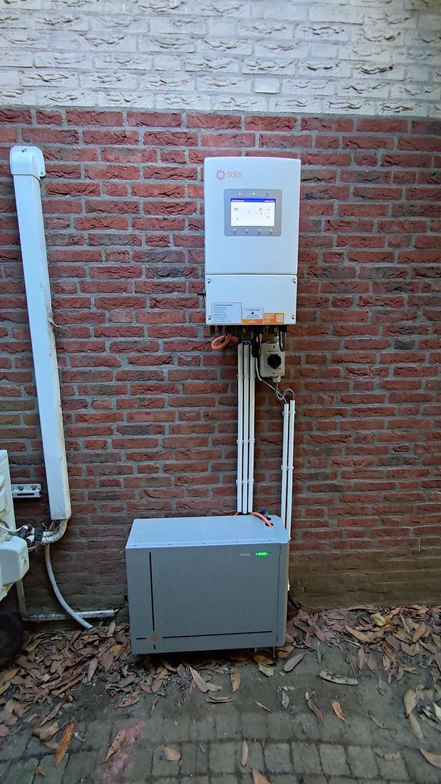 Een grijze accukast buiten tegen een bakstenen gevel, met de omvormer erboven